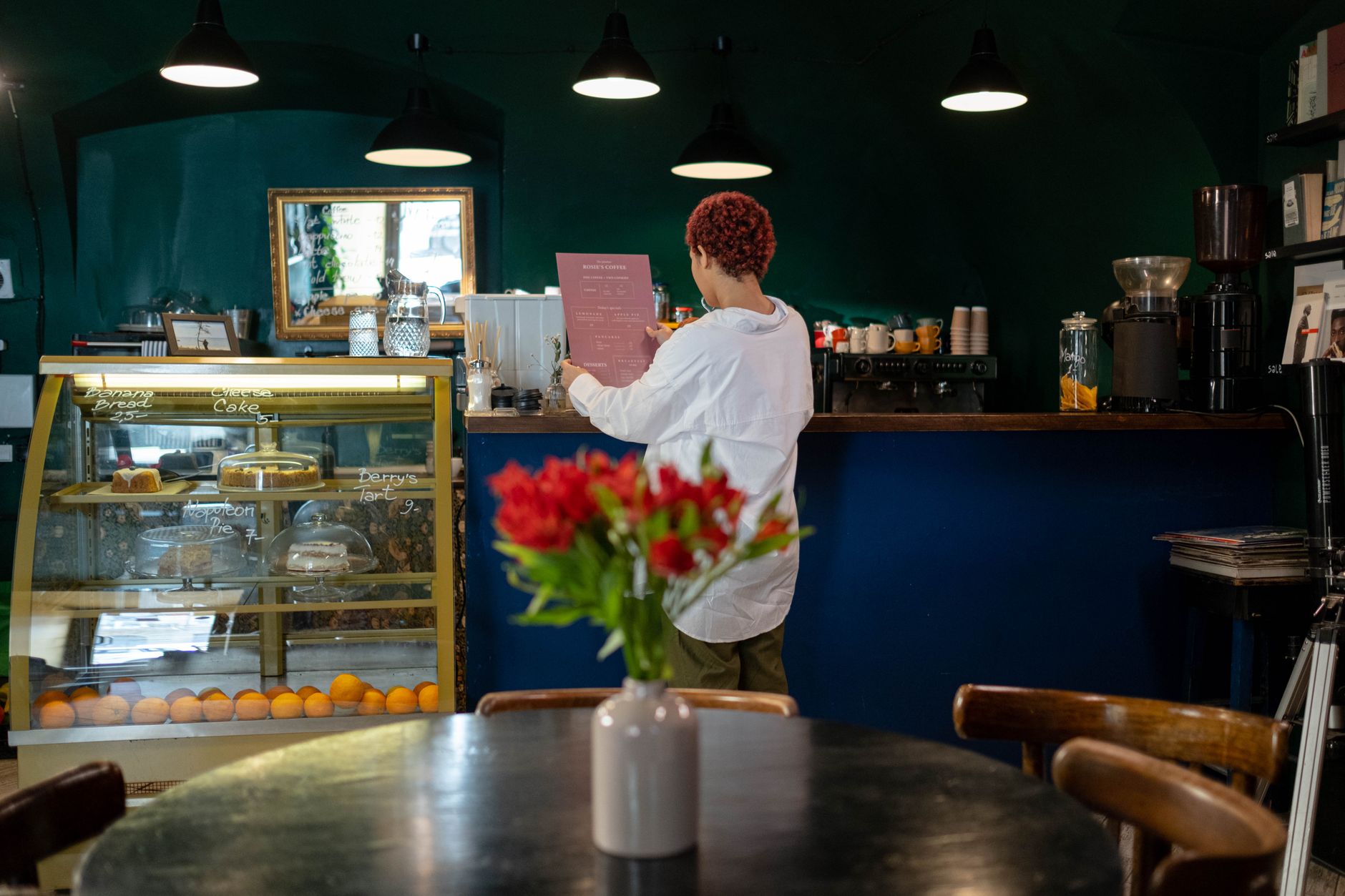 A barista reads an order in a cozy café with a pastry display and warm lighting.