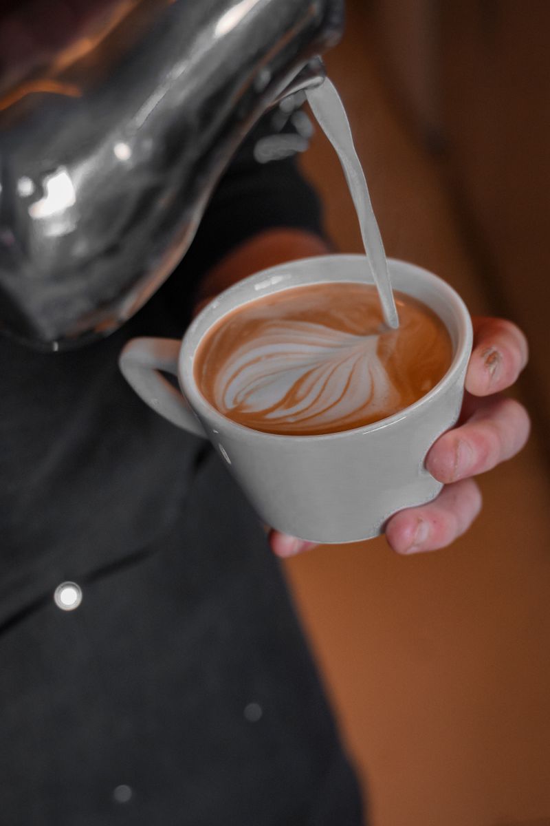 Close-up of hands pouring frothy milk into coffee cup creating intricate latte art.